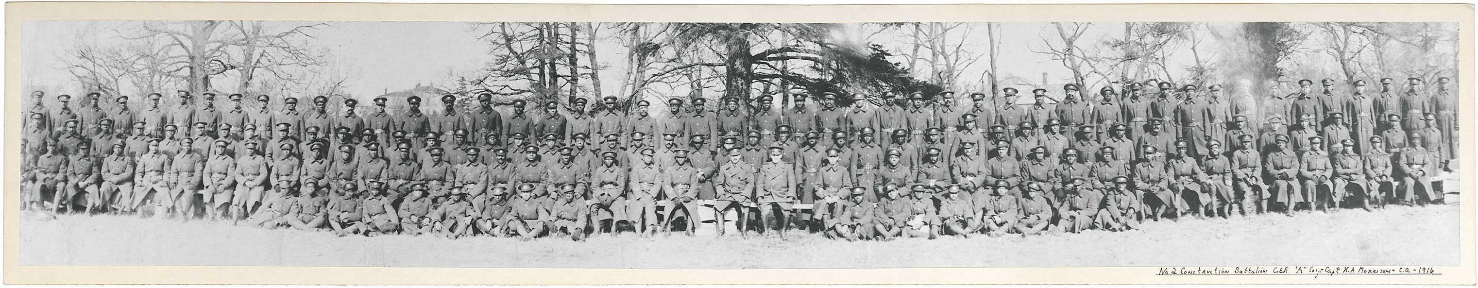 Photographie en noir et blanc – De nombreux hommes noirs sont assis pour une photo de groupe. La plupart portent de longs manteaux militaires. Des arbres et des toitures sont visibles à l’arrière-plan.
